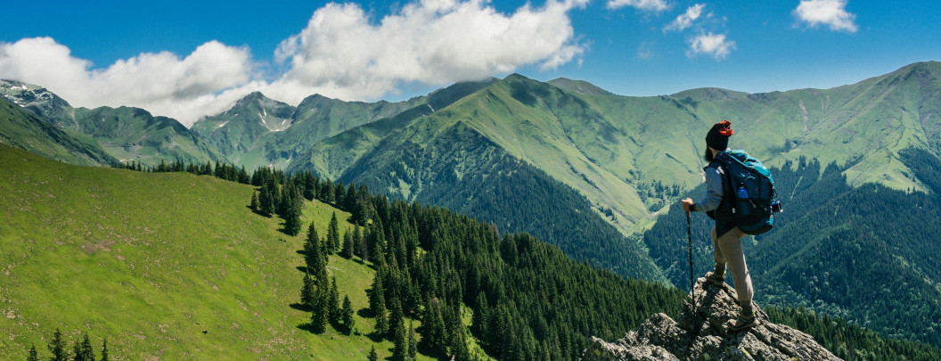 A person wearing a backpack stands on a rocky ledge overlooking a range of green mountains under a partly cloudy blue sky.