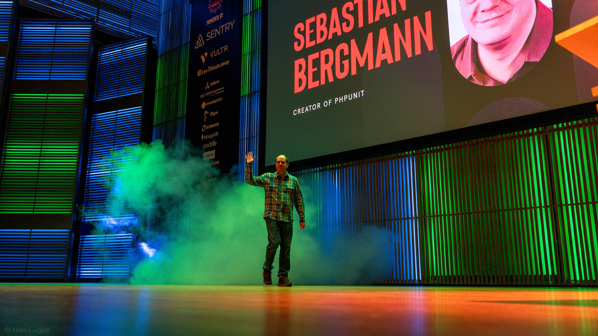 Sebastian Bergmann walks onto a stage through green and blue smoke, one hand raised in greeting, in front of a large screen that reads SEBASTIAN BERGMANN, CREATOR OF PHPUNIT next to a portrait of him.