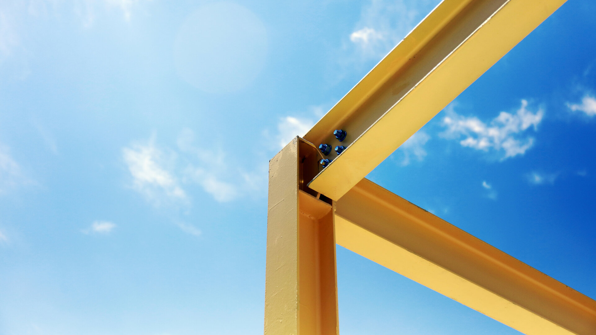 This image shows the bolted corner joint of a yellow steel frame, photographed from below against a blue sky with a few clouds. Two beams meet at a right angle above an upright column, held by four blue bolts. The structure continues past the edge of the frame in both directions. The imagery captures the essence of the article: the joint is not what anyone looks at, and everything built above it depends on whether it holds.