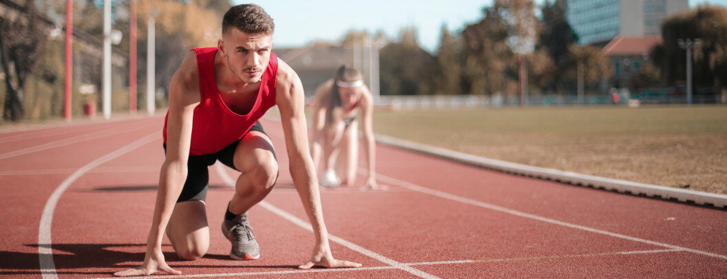 The image shows two people starting on a running track. In the foreground, a person in a red sports outfit is seen in the starting position for a sprint, ready to run. In the background, another person is also in the starting position. The image conveys a sporty and competitive moment on an outdoor athletics track in daylight, surrounded by trees and buildings.