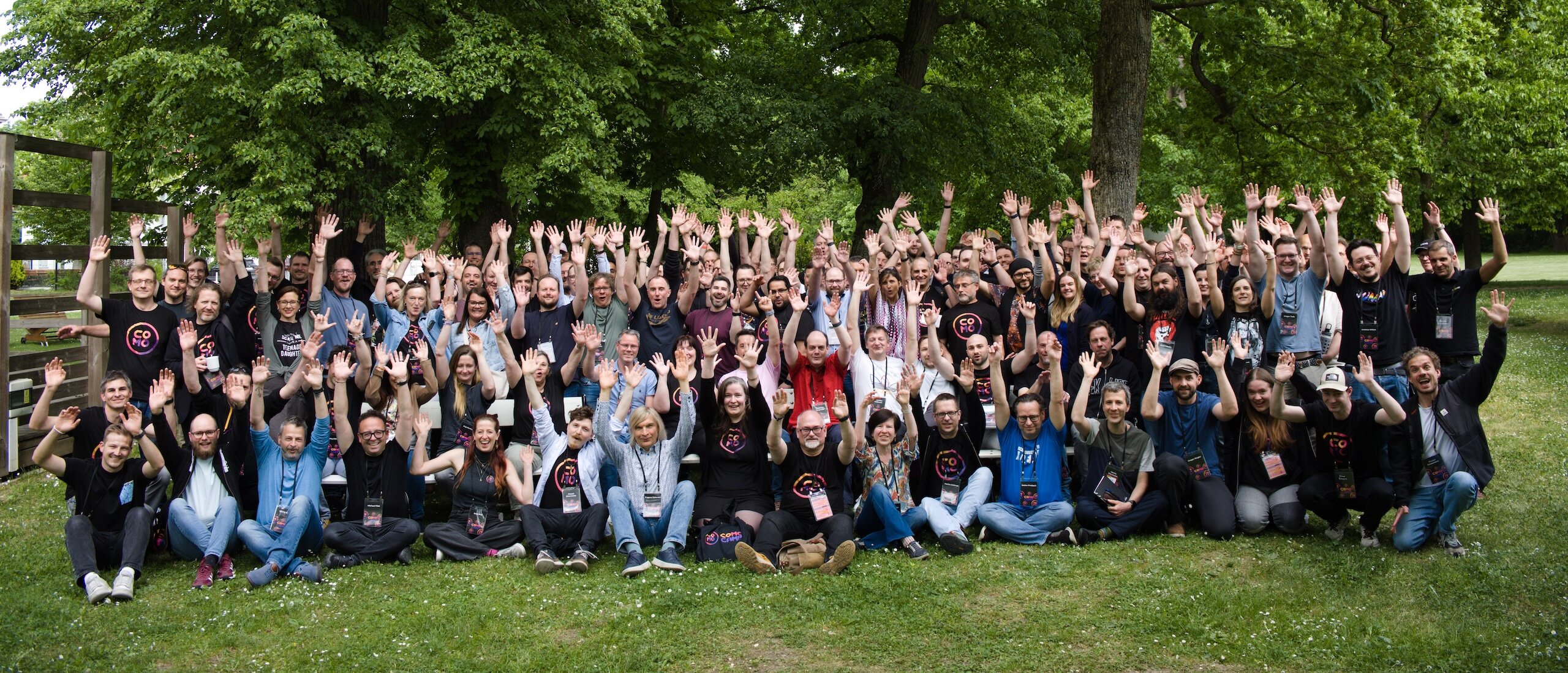 A group photo outdoors: the participants of ComoCamp 2026 stand and sit closely together on a meadow, with tall leafy trees in the background. The mood seems cheerful and warm; the rich green and the natural light give the image a summery, relaxed community atmosphere.