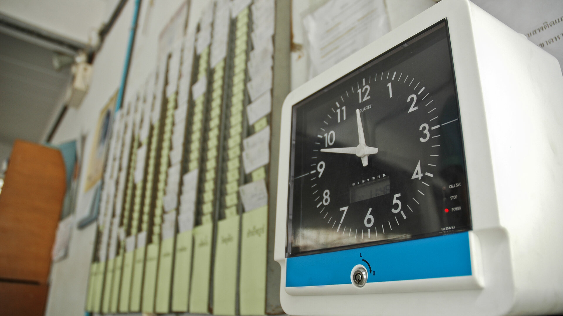 A white time clock with a large black dial stands in the foreground on a shelf, with a key lock set into its casing below the dial. Behind it, out of focus, rows of time cards fill their racks on the wall. A visual metaphor for a locked clock that nobody who clocks in can set.