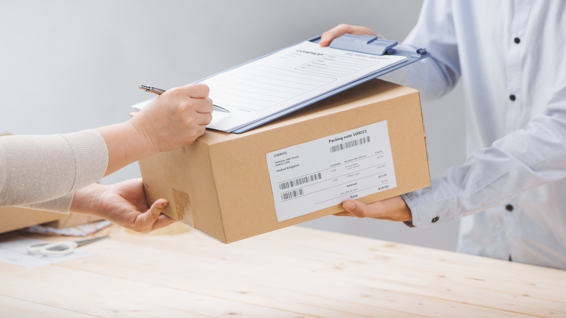 A sealed shipping box is handed across a counter, along with a clipboard holding a form. A packing note listing the enclosed items with quantities and prices is affixed to the box. A visual metaphor for a bill of materials that is created while packing and ships with the package instead of being guessed at afterwards.