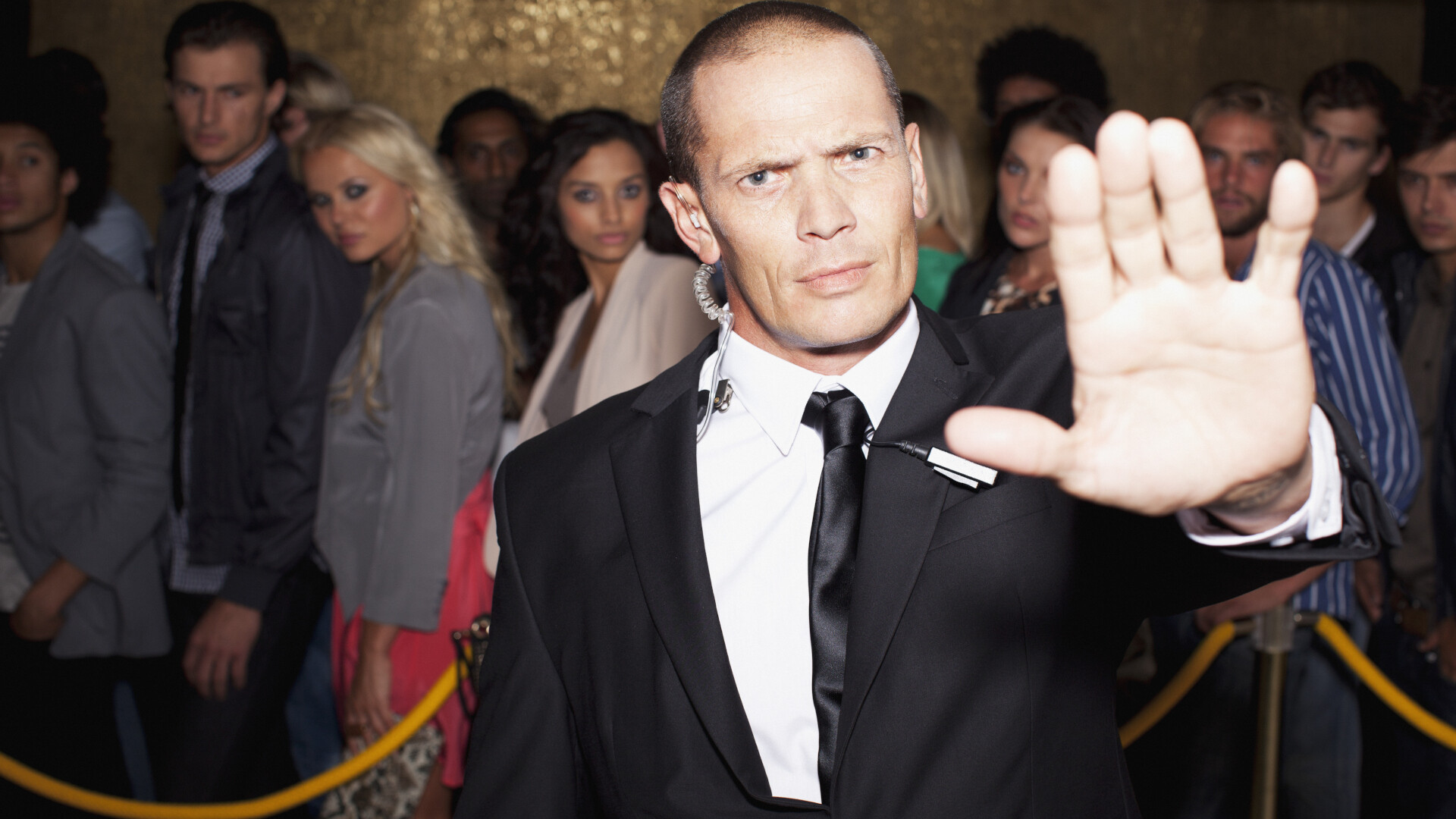 A bouncer in a black suit, earpiece in, hand raised in a firm "stop" gesture at the door of a club. Behind him, a queue of hopeful guests waits behind a golden velvet rope. A visual metaphor for a gatekeeper deciding who gets in and who does not.