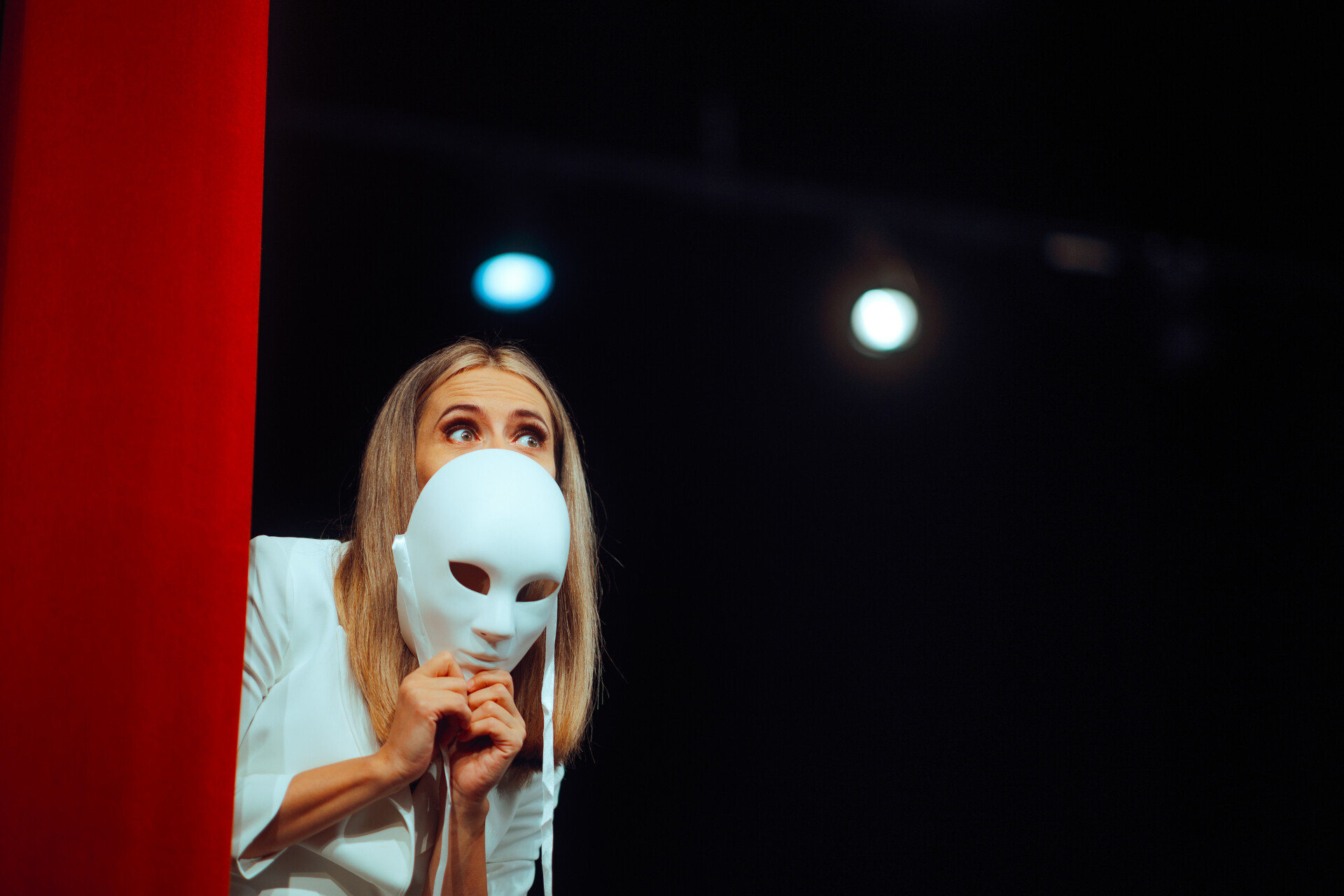 Dieses Bild zeigt eine Frau, die eine weiße Theatermaske vor ihrem Gesicht hält, vor einem dramatischen, dunklen Hintergrund. Links ist ein auffälliges rotes Feld zu sehen, und im Hintergrund gibt es eine stimmungsvolle Bühnenbeleuchtung mit blauen und weißen Kugeln. Ihre Augen schauen über die Maske hinweg, was auf einen Moment der Selbstreflexion oder Erkenntnis hindeutet, mit einem Ausdruck von Neugier und leichter Besorgnis. Die Theaterkulisse und die professionelle Inszenierung betonen den performativen Charakter des Augenblicks.
