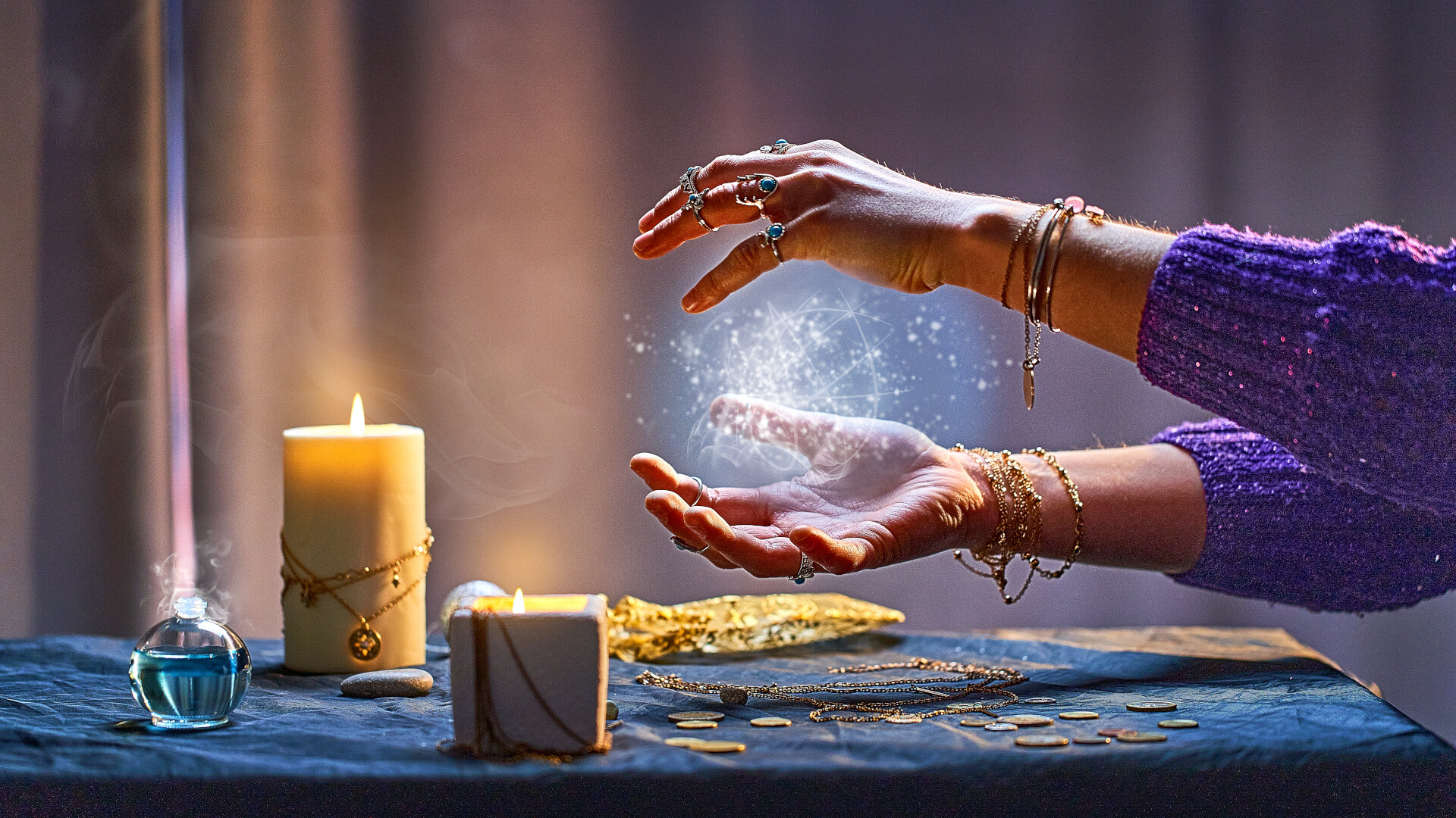 The image shows two hands that seem to be forming a glowing light that appears magical. The person is wearing several rings and bracelets and has their arm tucked into a purple jumper. In the foreground is a table covered with a dark cloth, on which lie candles, coins, chains and a small bottle containing a blue-green liquid. The warmly lit scene evokes a spiritual or mystical atmosphere, reminiscent of a ritual or fortune telling.