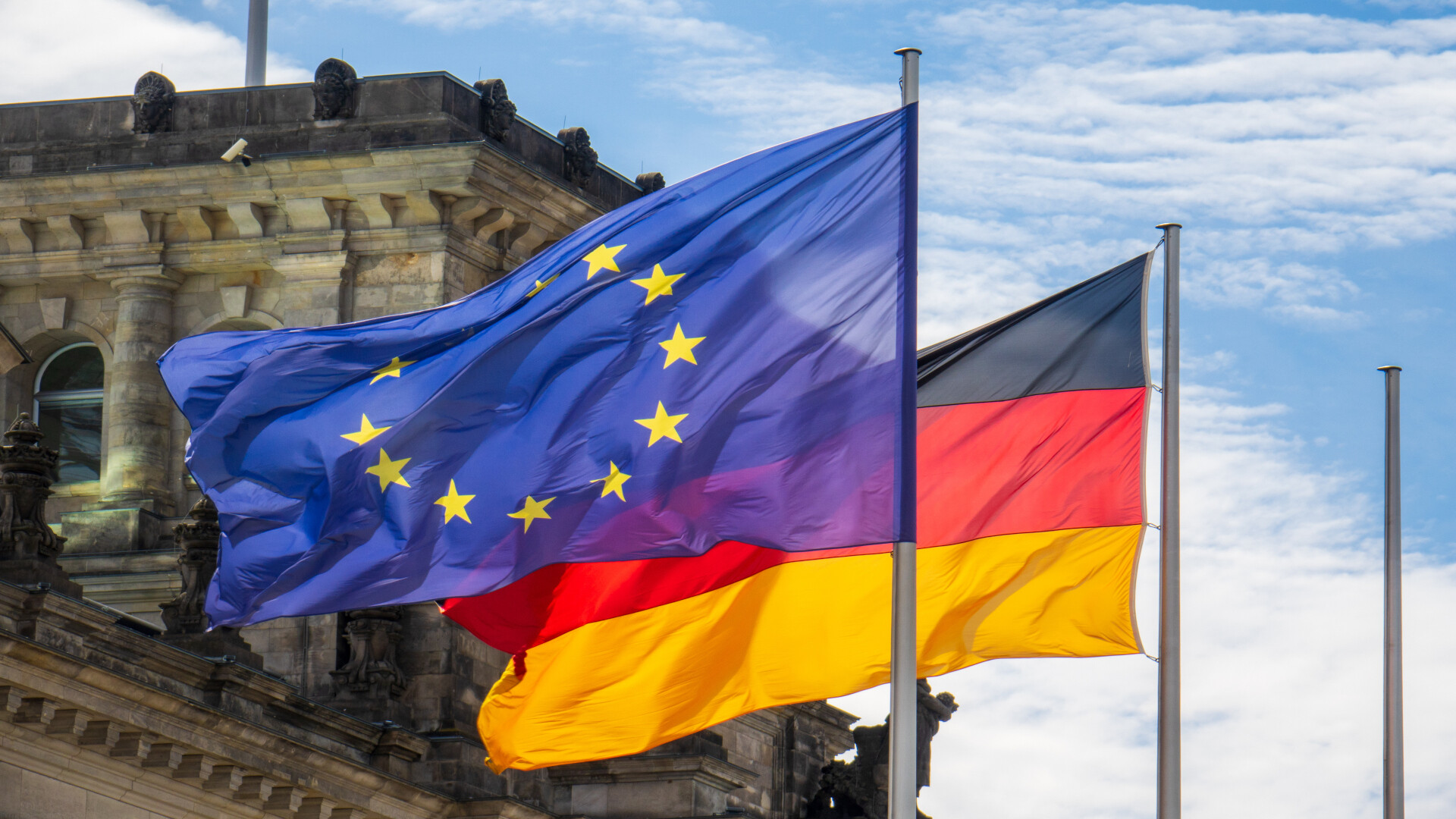 The flag of the European Union and the German national flag flying side by side in front of the Reichstag building in Berlin.