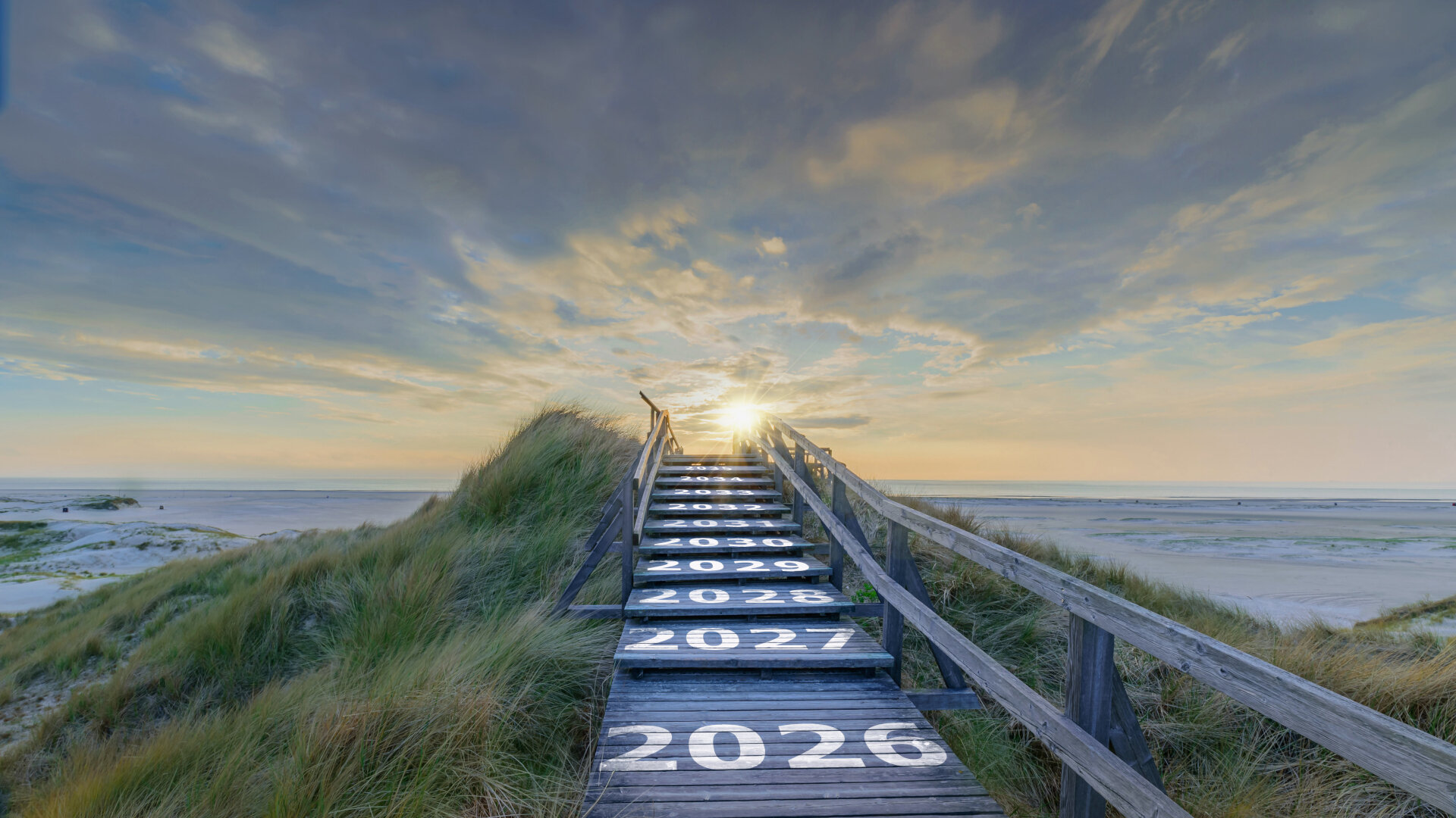 Das Bild zeigt eine hölzerne Treppe, die durch Dünen zum Strand führt und in Richtung eines Sonnenaufgangs oder Sonnenuntergangs verläuft. Auf den einzelnen Stufen sind Jahreszahlen von 2026 bis 2035 groß und weiß aufgemalt, als würden sie den Weg in die Zukunft symbolisieren. Am Ende der Treppe, wo das Licht am stärksten ist, scheint die Reise in die kommenden Jahre zu führen, während der breite Himmel und das ruhige Meer im Hintergrund eine friedliche, weite Stimmung vermitteln.