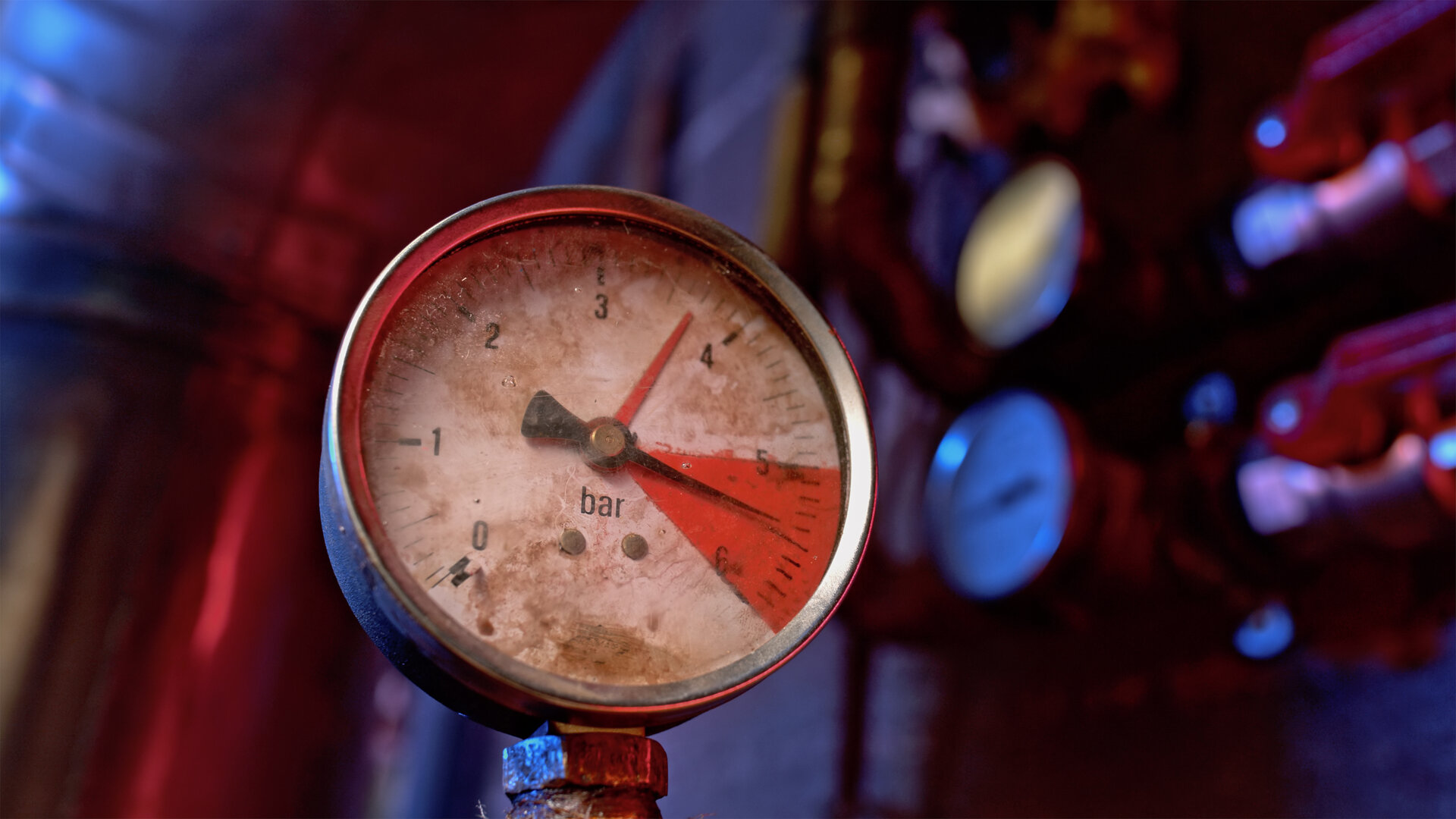 This image shows an old, grimy pressure gauge in the foreground of a machine room. Its black needle stands between five and six bar, in the middle of the section of the scale marked in red. Blurred pipes, valves and further gauges in red and blue light fill the background. The imagery captures the essence of the article: the problem sat in the red zone for a long time, and whether the system holds depends on the few people who maintain it.
