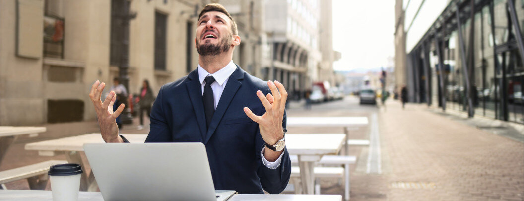 In the picture, a man in a suit is sitting outdoors at a table in front of a laptop. He has raised both hands in frustration and is looking up. His facial expression appears desperate or annoyed. The scene is set on an empty street with benches and buildings in the background. There is also a coffee cup on the table in front of him.