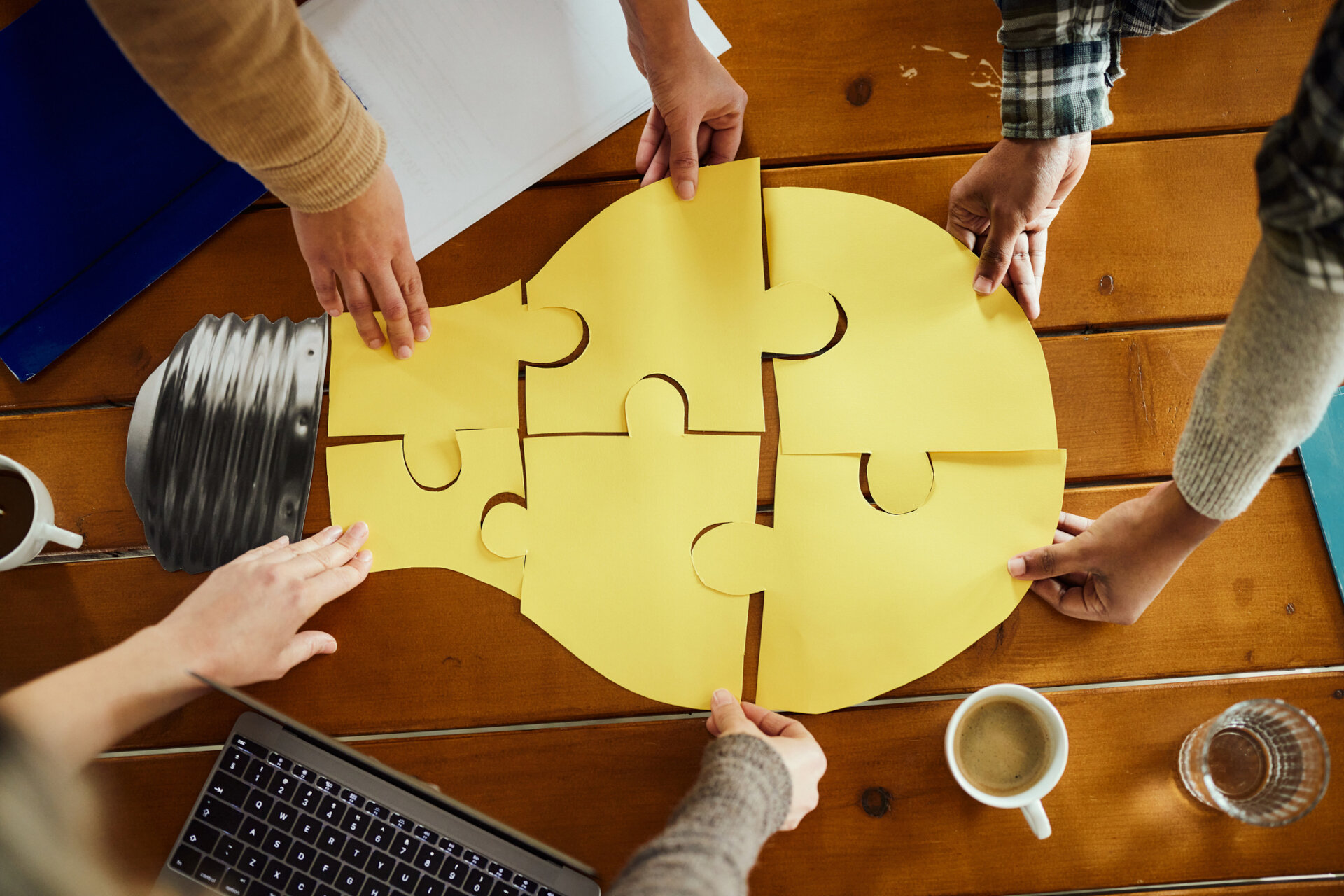 The image shows several people working together to assemble a puzzle made up of yellow pieces that form the shape of a light bulb. The light bulb typically symbolises an idea or innovation. The scene takes place on a wooden table, which also holds a cup of coffee, a glass of water, an open laptop and some documents. The people