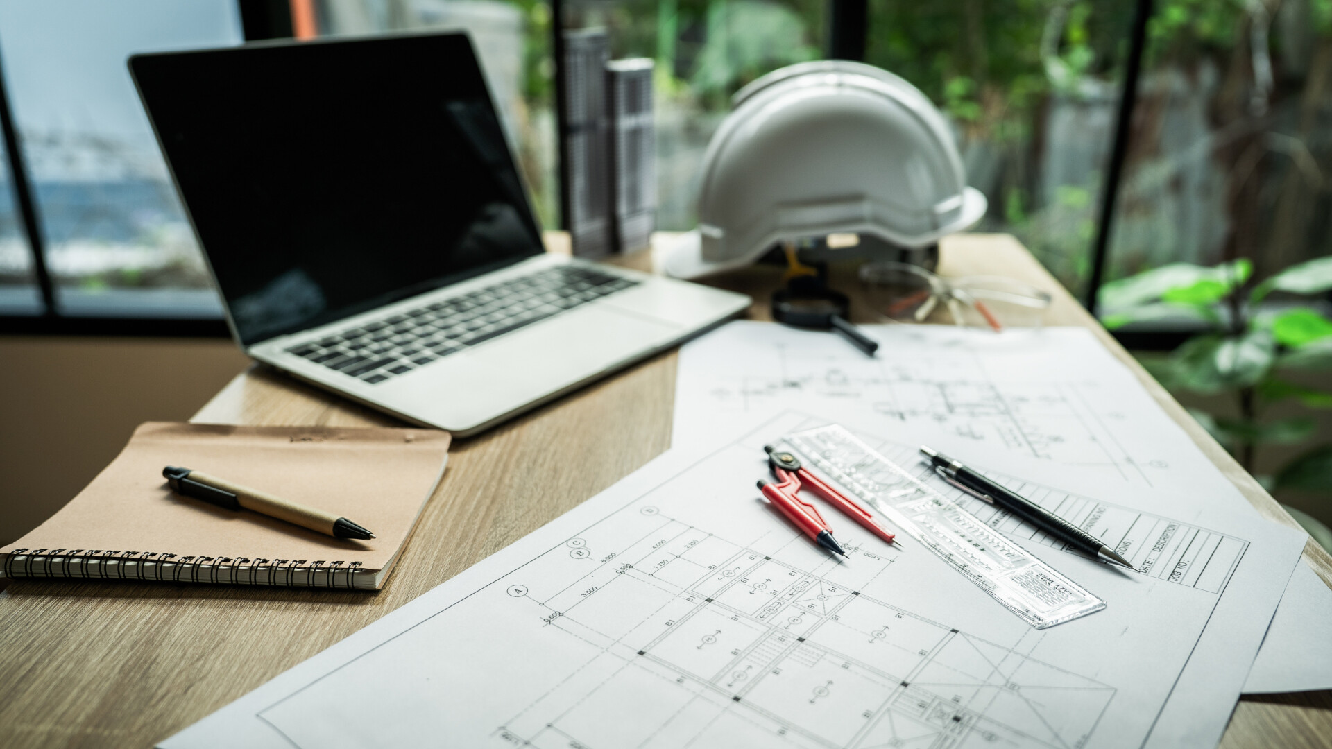 This photograph captures a professional workspace set up outdoors against a backdrop of a natural garden. The scene showcases an organised desk arrangement on a light wooden surface, bathed in daylight.
