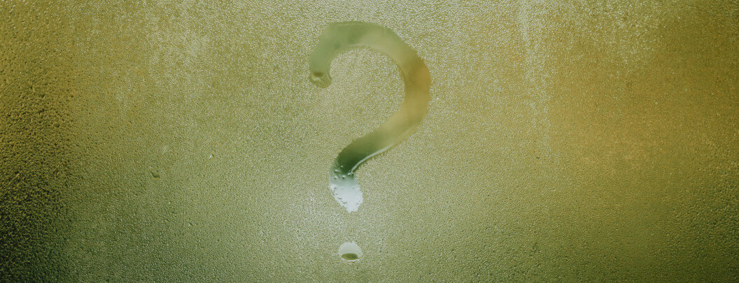 The image shows a glass surface covered with fine water droplets or condensation. A large question mark has been painted or wiped onto this fogged-up glass. The background appears yellowish to greenish and blurred, so that the question mark clearly stands out as the central motif.