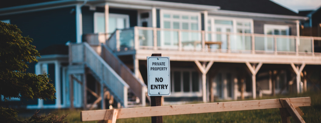 A sign reading "Private Property No Entry" stands in front of a large modern house with a wooden deck and glass railings in the background.