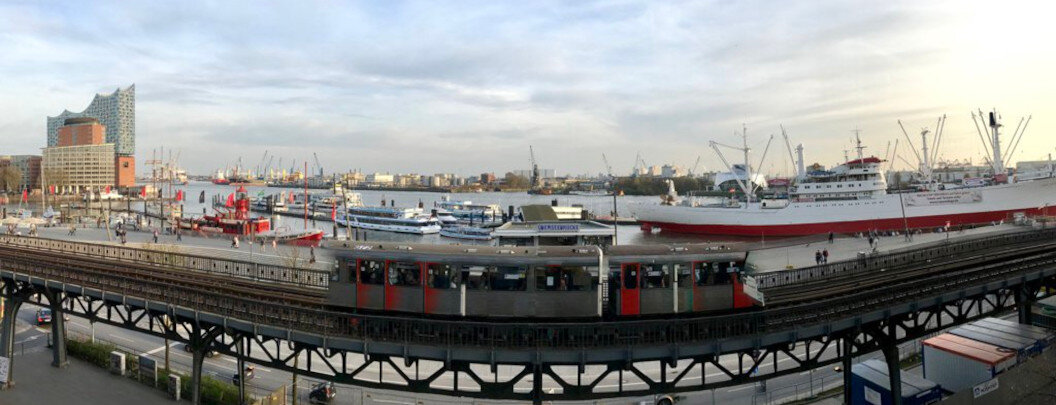 The image shows a panoramic view of a harbour with a train on an elevated railway bridge in the foreground. On the left is a modern building with a striking glass façade, which is the Elbphilharmonie concert hall in Hamburg. In the background, several ships and cranes can be seen, indicating port activity. The weather is cloudy but bright, suggesting late afternoon or early evening. The scene appears both calm and industrial.