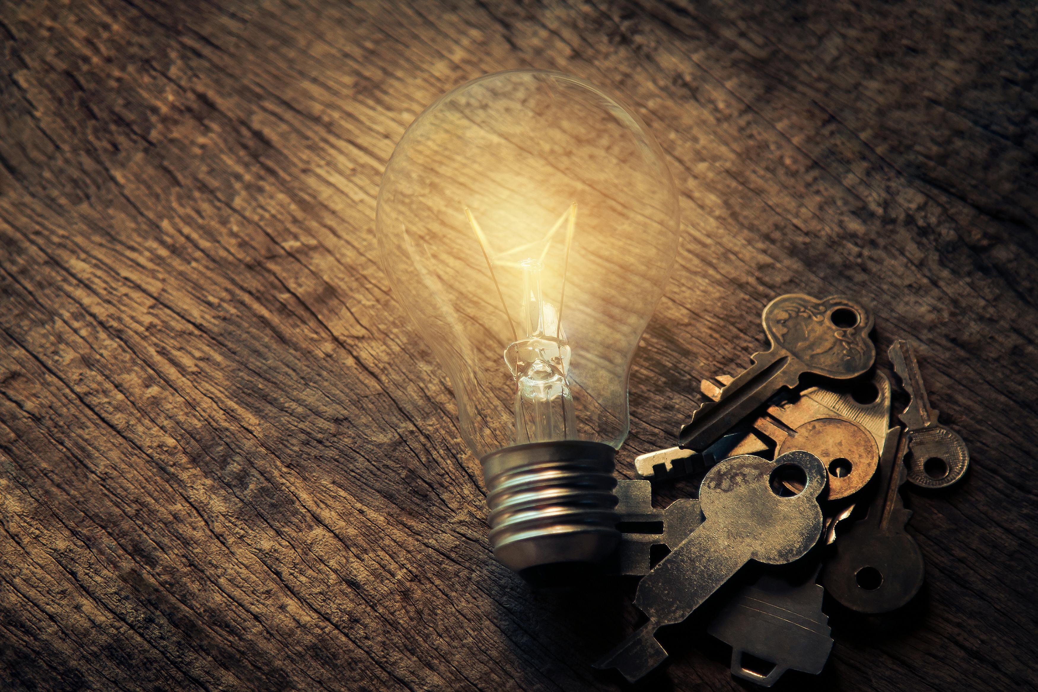 The picture shows a light bulb lying on a wooden surface. The light bulb is lit and creates a warm atmosphere. Right next to the light bulb is a small pile of various old metal keys. The interplay between the glowing light bulb and the keys symbolically conveys the idea that ideas (light bulb) can be the key (bunch of keys) to new possibilities or solutions.