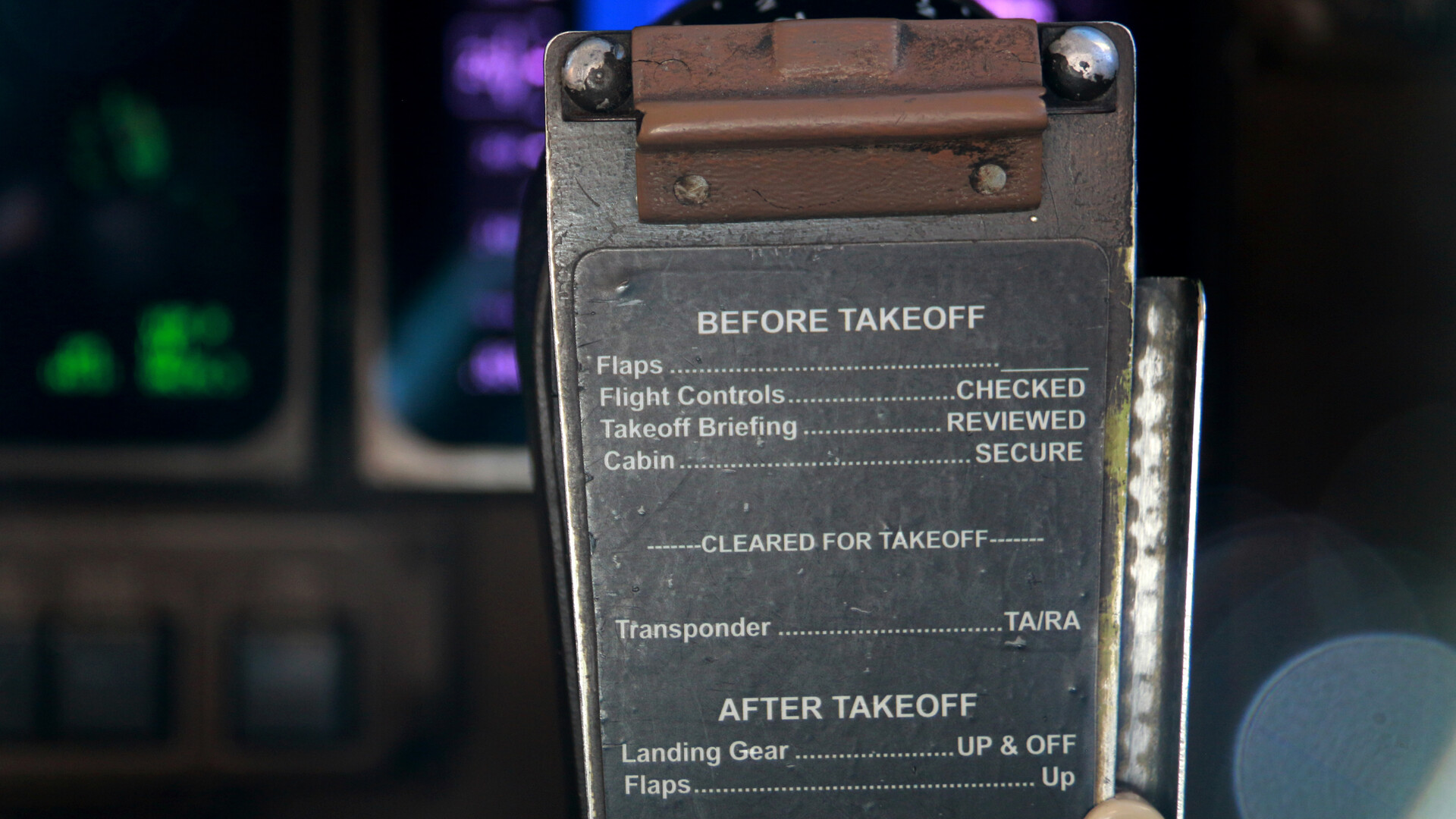 A worn metal checklist card hangs in the cockpit of an aircraft in front of dimly glowing flight instruments. The card is engraved with a list titled BEFORE TAKEOFF, whose items read Flight Controls CHECKED, Takeoff Briefing REVIEWED and Cabin SECURE, followed by a dashed line reading CLEARED FOR TAKEOFF and the beginning of an AFTER TAKEOFF section.