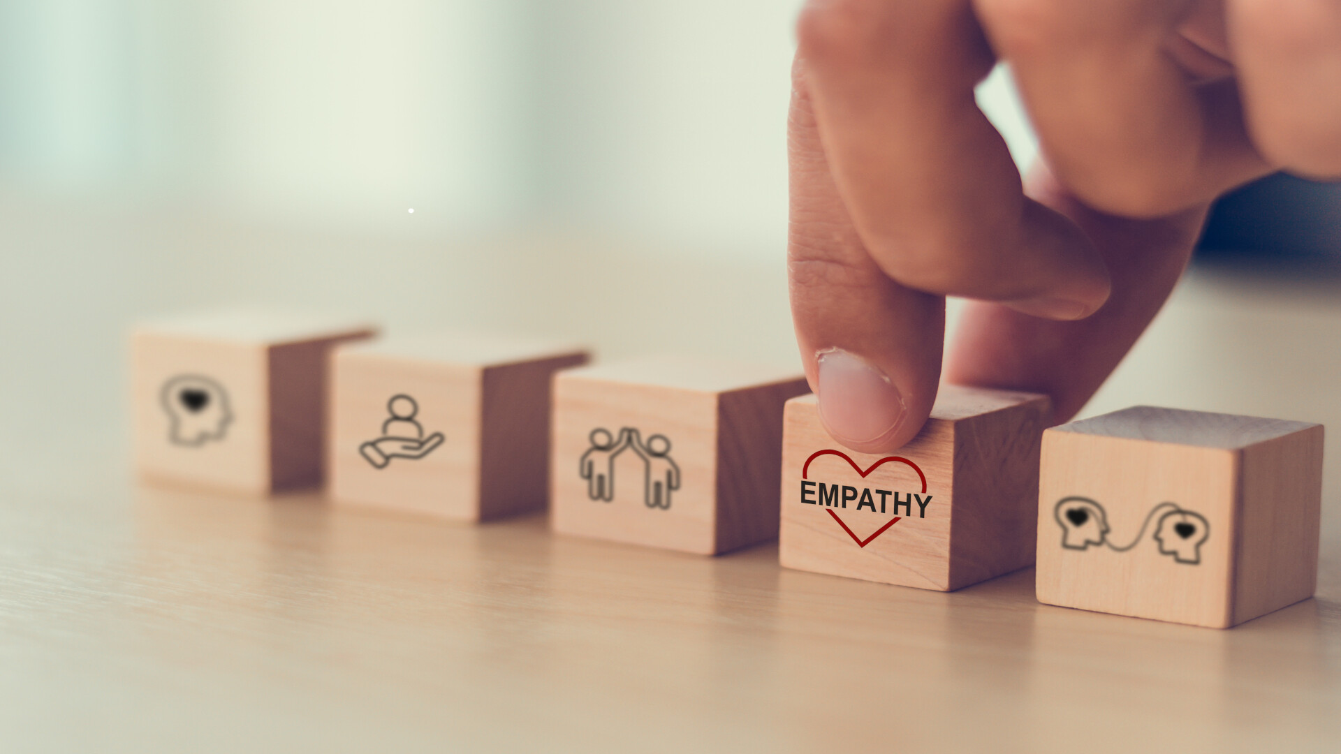 The image shows five wooden blocks with symbols representing interpersonal skills. A hand deliberately highlights the cube with the red heart and the inscription "EMPATHY". The warm, calm atmosphere emphasises empathy as a technical skill in software development.