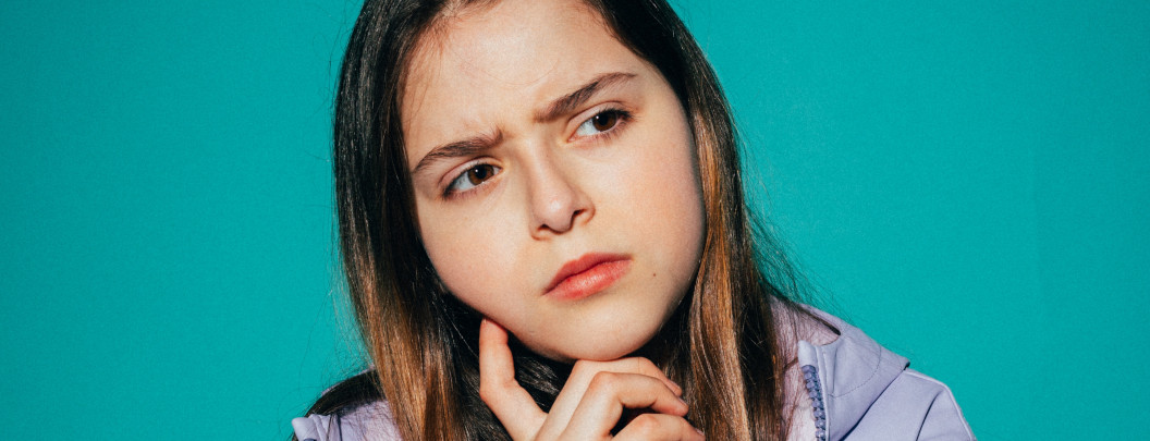 A young person with long brown hair looks thoughtful and slightly concerned, resting their chin on their hand against a teal background.