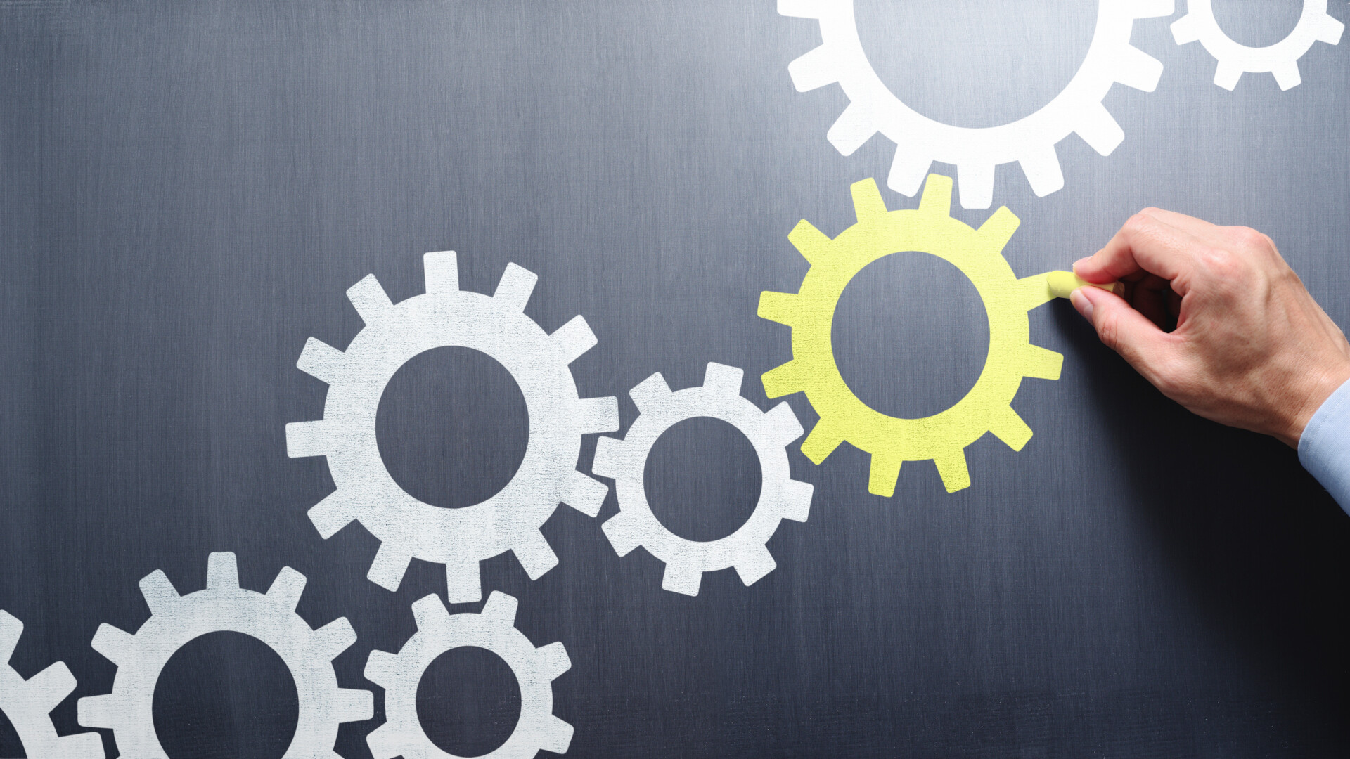 Das Bild zeigt mehrere ineinandergreifende Zahnräder in Weiß und Gelb auf einer dunklen Tafeloberfläche. Eine Hand ergänzt oder hebt gerade eines der gelben Zahnräder hervor. Diese Hand kann als Sinnbild für den Entwickler gelesen werden, der gezielt neue, typensichere Komponenten in ein bestehendes System einfügt, ohne den Mechanismus zu stören.