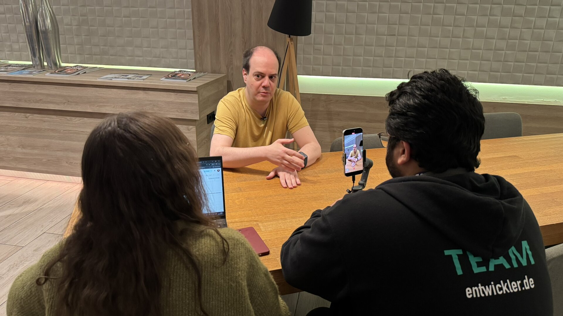 Sebastian Bergmann, in a yellow T-shirt with a lapel microphone, sits at a long wooden table and explains something with one hand raised. In the foreground, two people sit with their backs to the camera: on the left, Ines Chargui, with long dark hair and a green knitted jumper, sits in front of an open laptop; on the right, someone in a black hoodie printed with “TEAM entwickler.de” films the interview with a smartphone on a gimbal, its screen showing Sebastian. In the background, a floor lamp with a black shade and a low shelf with magazines and silver vases stand in front of a backlit wall.