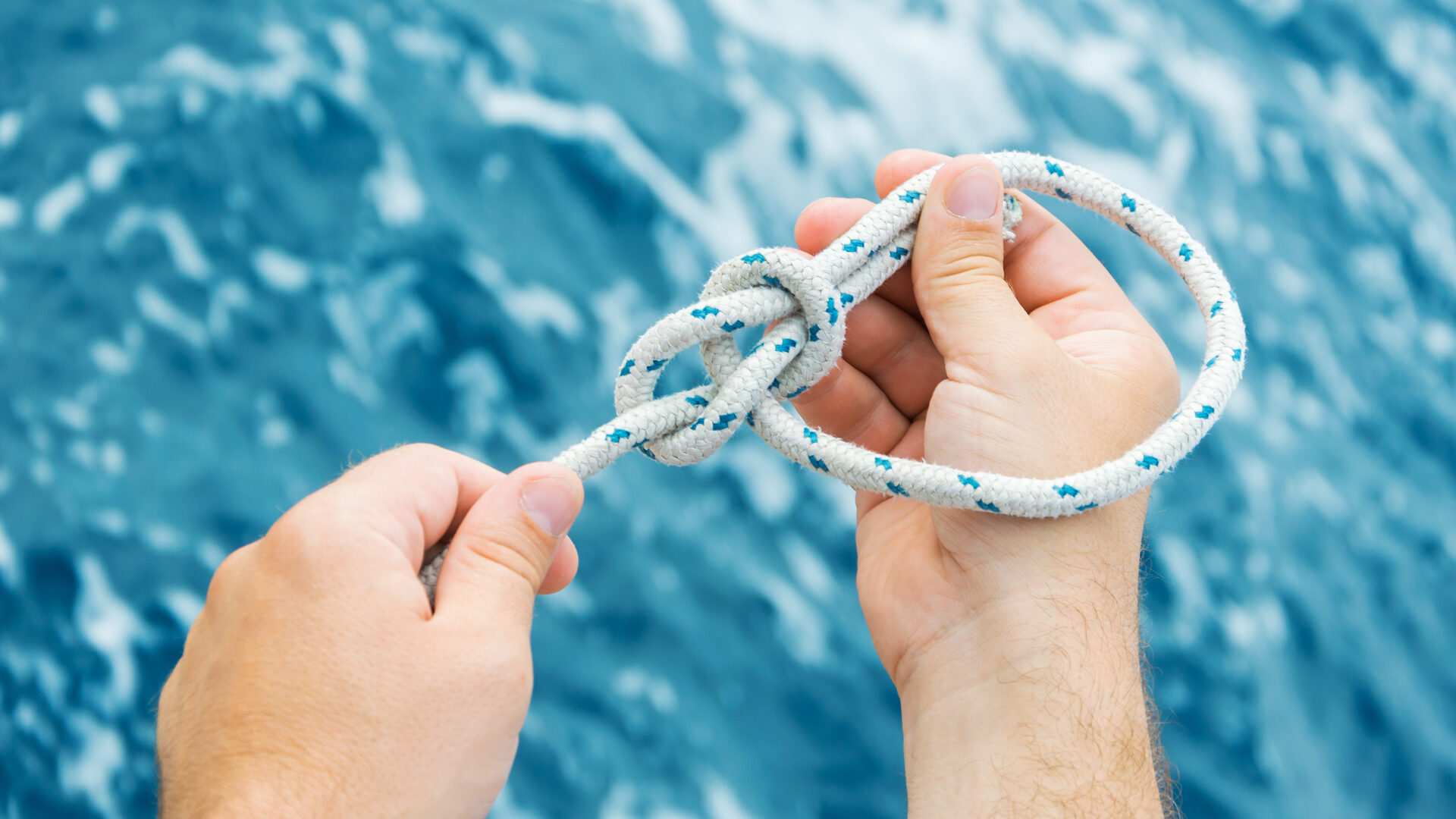 Die Hände einer Person knüpfen einen Palstek in ein Segeltau, im Hintergrund das Wasser. Ein Sinnbild für Verifikation: Vertrauen in einen Knoten erbt sich nicht von der Person, die ihn geknüpft hat. Wer sich darauf verlassen will, knüpft ihn selbst oder prüft ihn.