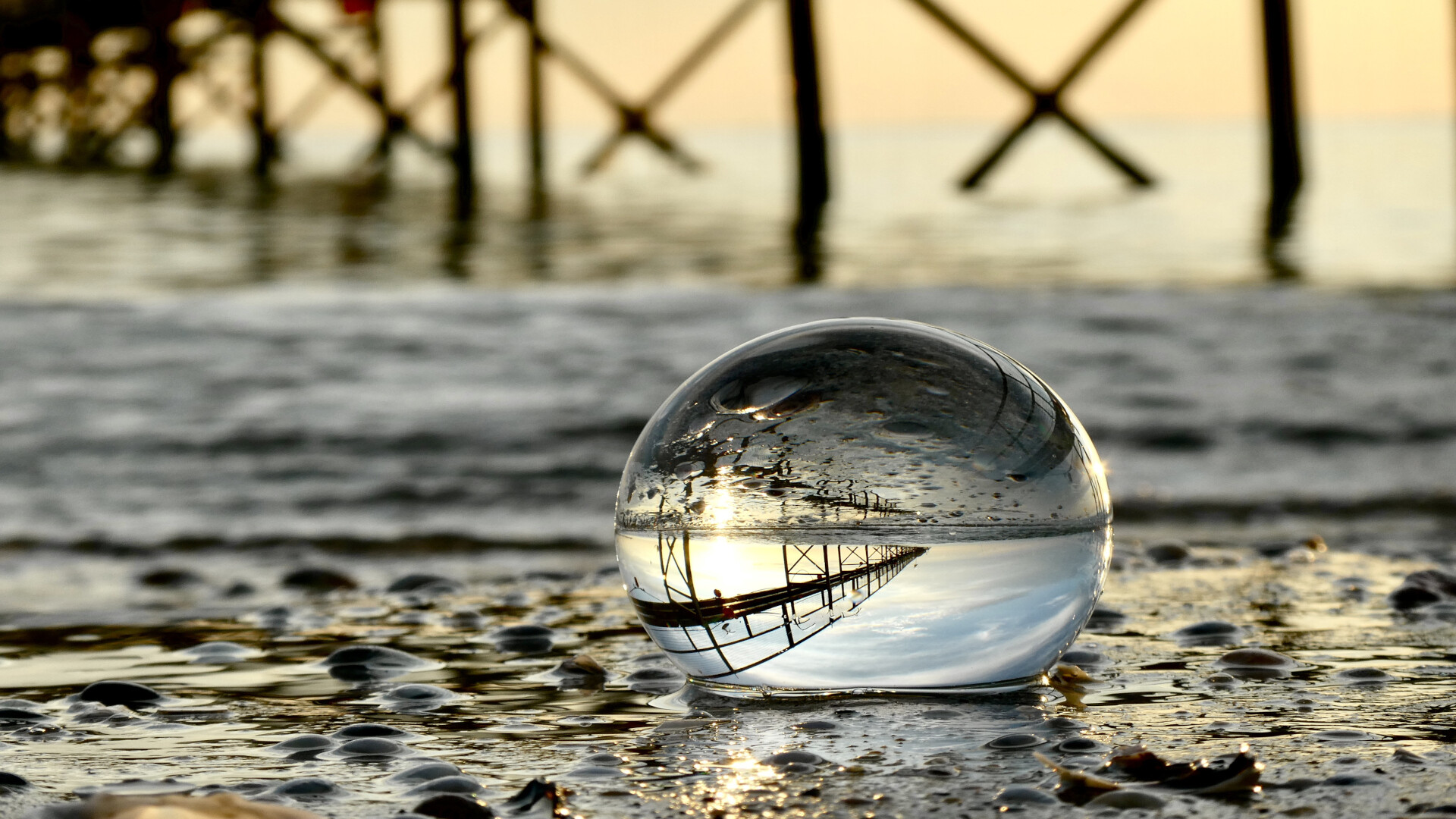 Glass sphere resting on wet pebbles at the shoreline, reflecting a pier and sunset, symbolizing a shift in perspective and bringing clarity to complex testing practices.