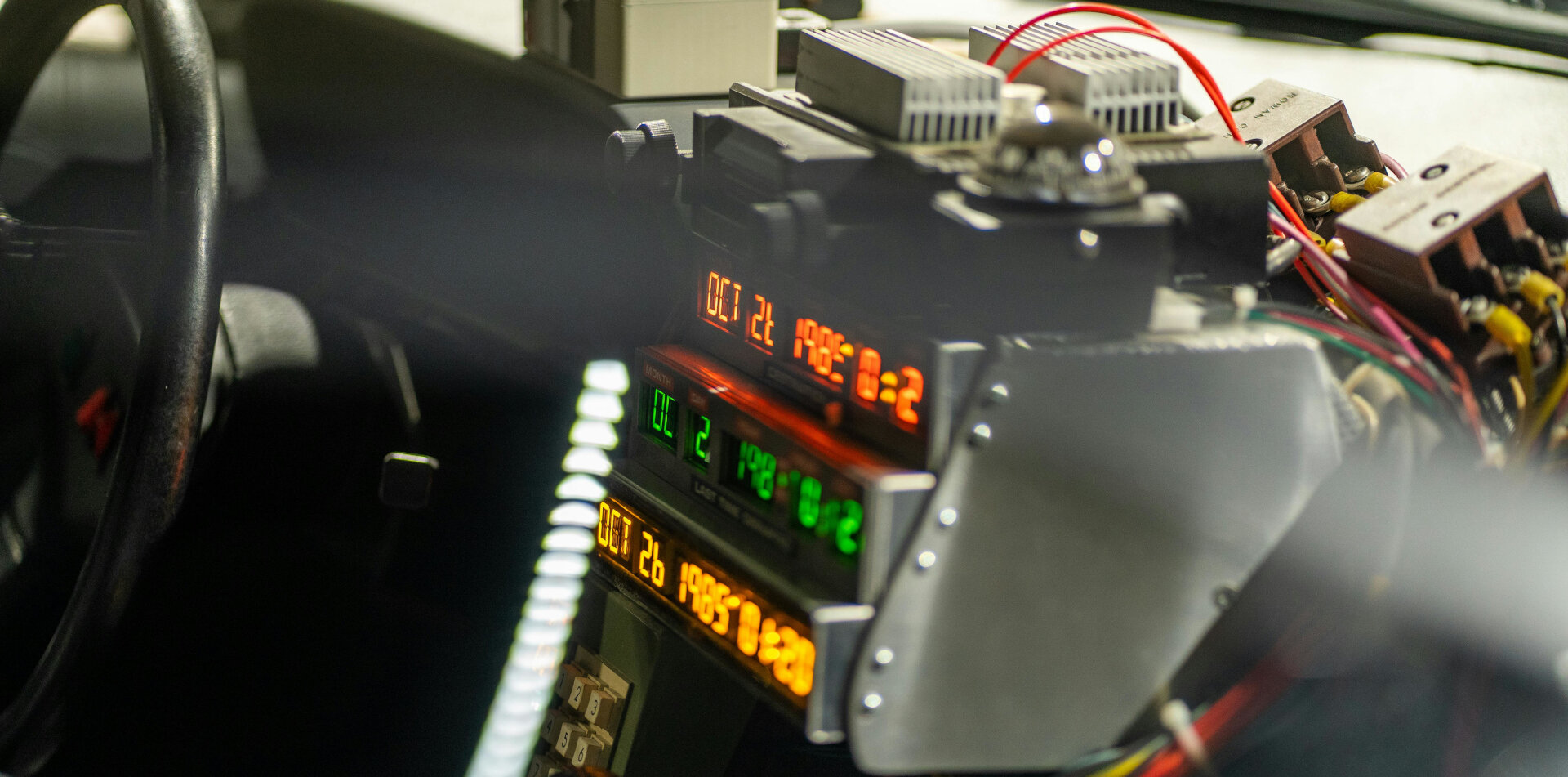 The image shows the interior of a vehicle with a futuristic-looking control panel consisting of several orange and green seven-segment displays arranged one above the other, showing the date and time, flanked by metal plates, switches, cables and connectors, creating the impression of a time travel console.