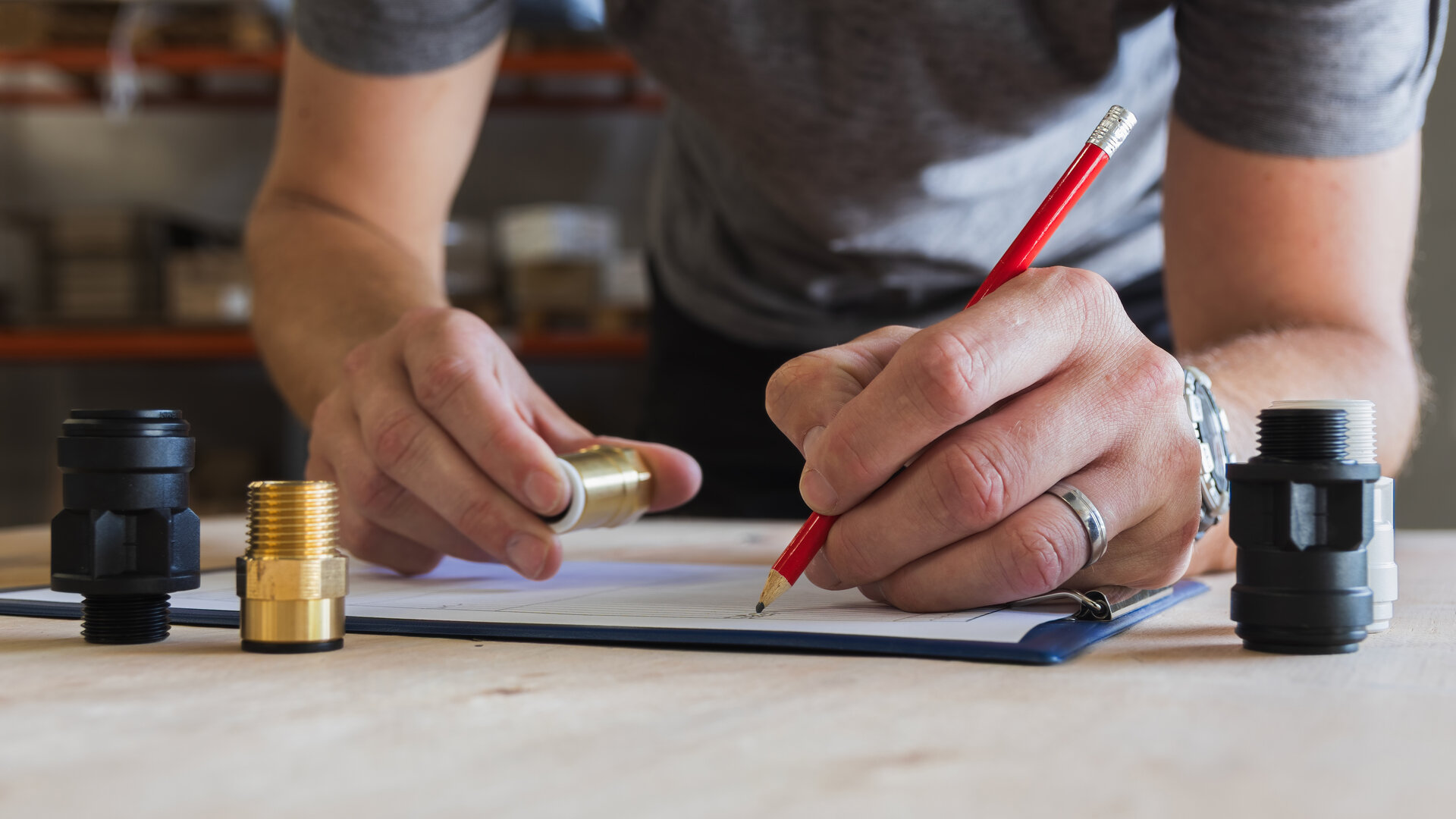 Die Hände einer Person an einer Werkbank: Die eine hält ein Fitting aus Messing, die andere schreibt mit einem roten Bleistift auf ein Klemmbrett, daneben stehen weitere Fittings. Ein Sinnbild für Verifikation: Wer wissen will, ob ein Teil stimmt, muss es prüfen und festhalten, was dabei zu beobachten war.