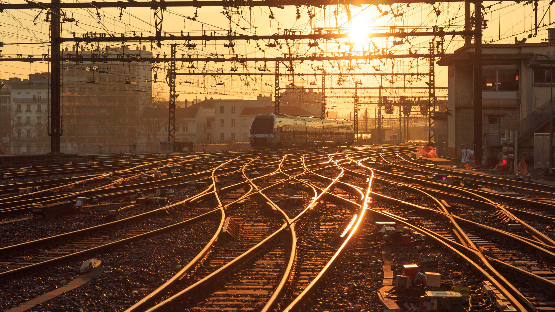 The approach to a station, backlit by a low sun. Dozens of rails branch apart over points and merge again, the railheads glowing gold. A railcar waits in the background under a dense web of overhead wires. A visual metaphor for the central question of every interpreter: how does execution get from one instruction to the next as quickly as possible?