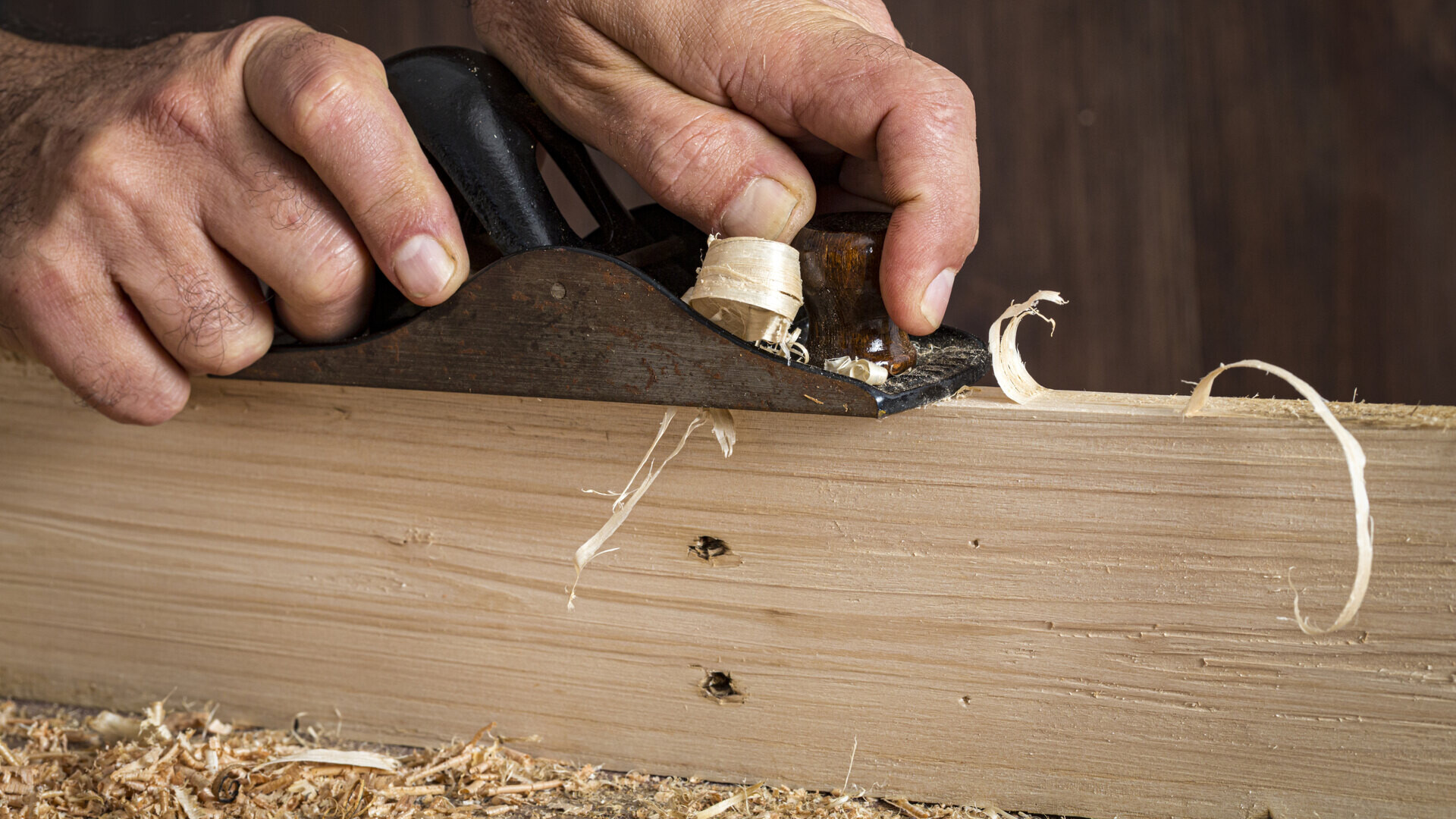 Two hands guiding a hand plane along a light-coloured board. A shaving runs out of the plane, a second one curls up on the wood in front of the blade. A heap of shavings lies below the board. The board itself is smooth and shows its grain; nothing has been added to it.