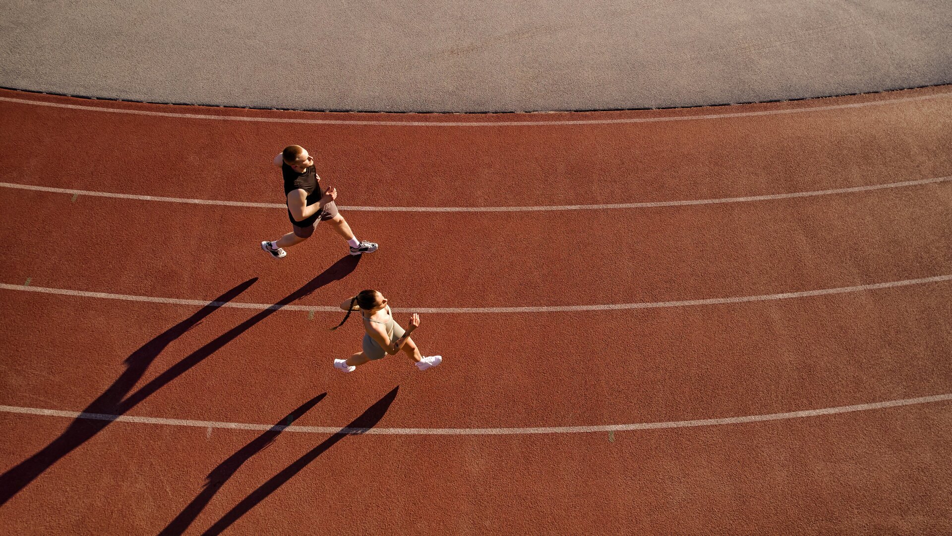 Zwei Personen laufen aus der Vogelperspektive über die rote Bahn eines Stadions, versetzt zueinander in benachbarten Bahnen. Die tiefstehende Sonne zieht ihre Schatten weit über den Belag, die weißen Linien durchziehen das Bild im immer gleichen Abstand. Ein Sinnbild dafür, dieselbe Strecke unter gleichen Bedingungen wieder und wieder zu laufen, bis das Muster sichtbar wird.