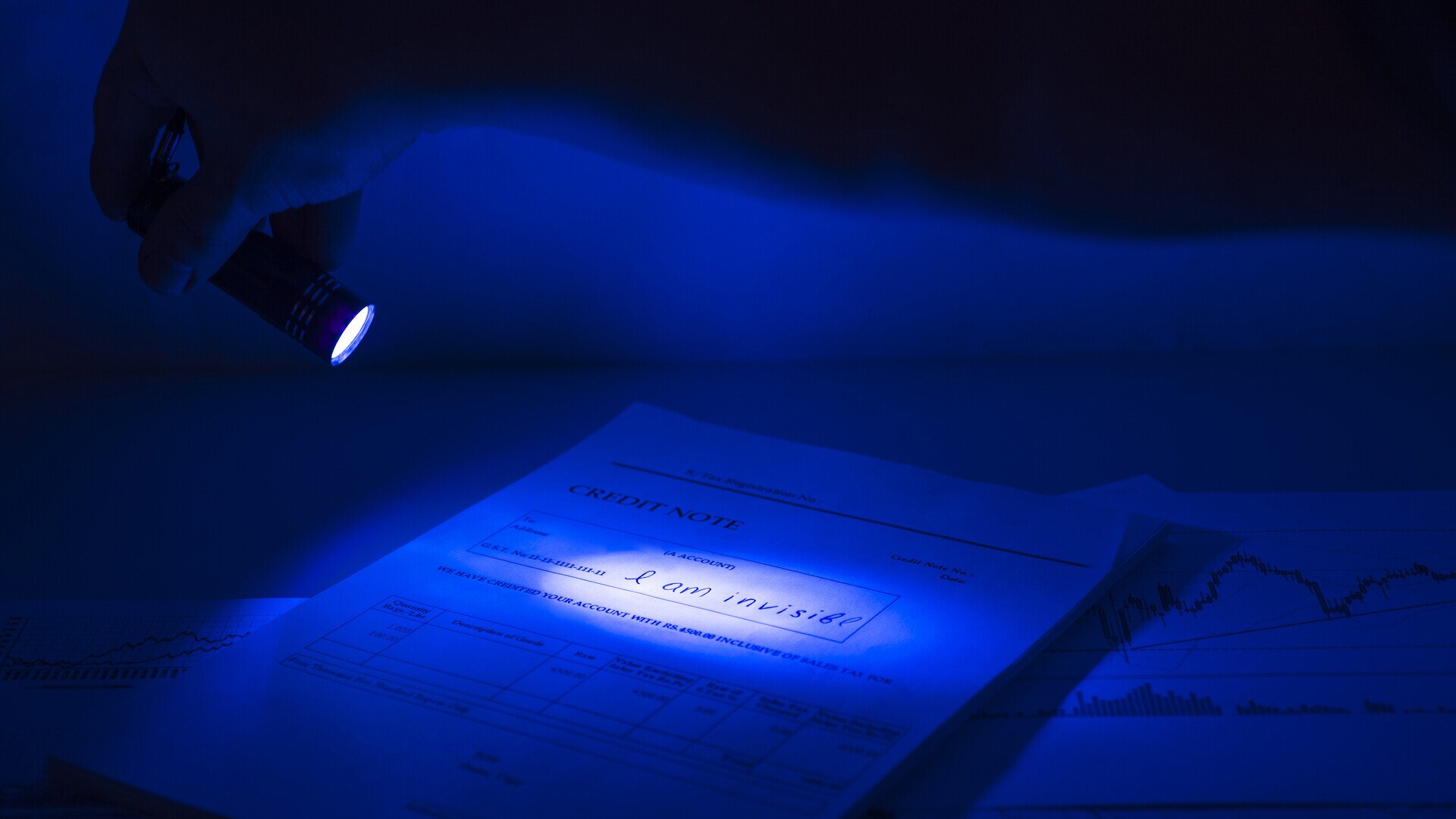 In a dark room, a hand holds a small ultraviolet torch over a printed form titled CREDIT NOTE. Where the beam falls on one of its fields, a handwritten line appears that the form does not show in ordinary light: I am invisible. More sheets with printed charts lie around it on the desk, barely visible in the blue light.