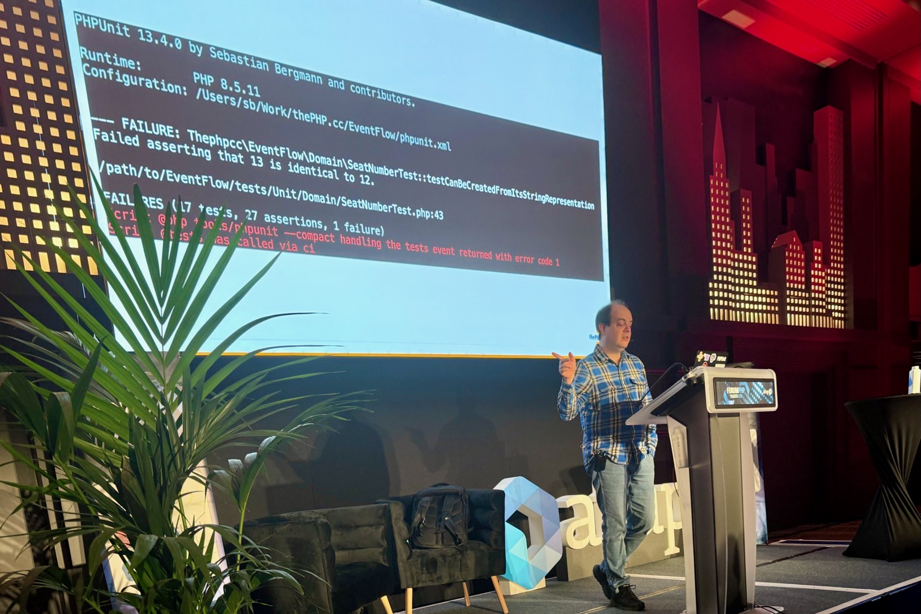 Sebastian Bergmann stands next to the lectern at Forum PHP 2026 and gestures towards the screen behind him, which shows the output of a failed PHPUnit test run.
